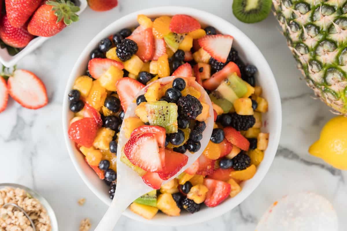 A bowl of mixed fruit salad with strawberries, blueberries, kiwi, blackberries, pineapple, and orange pieces; a spoon holds a portion above the bowl.