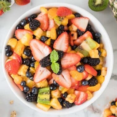 A bowl of mixed fruit salad with strawberries, kiwi, blueberries, blackberries, oranges, and pineapple, garnished with mint leaves.