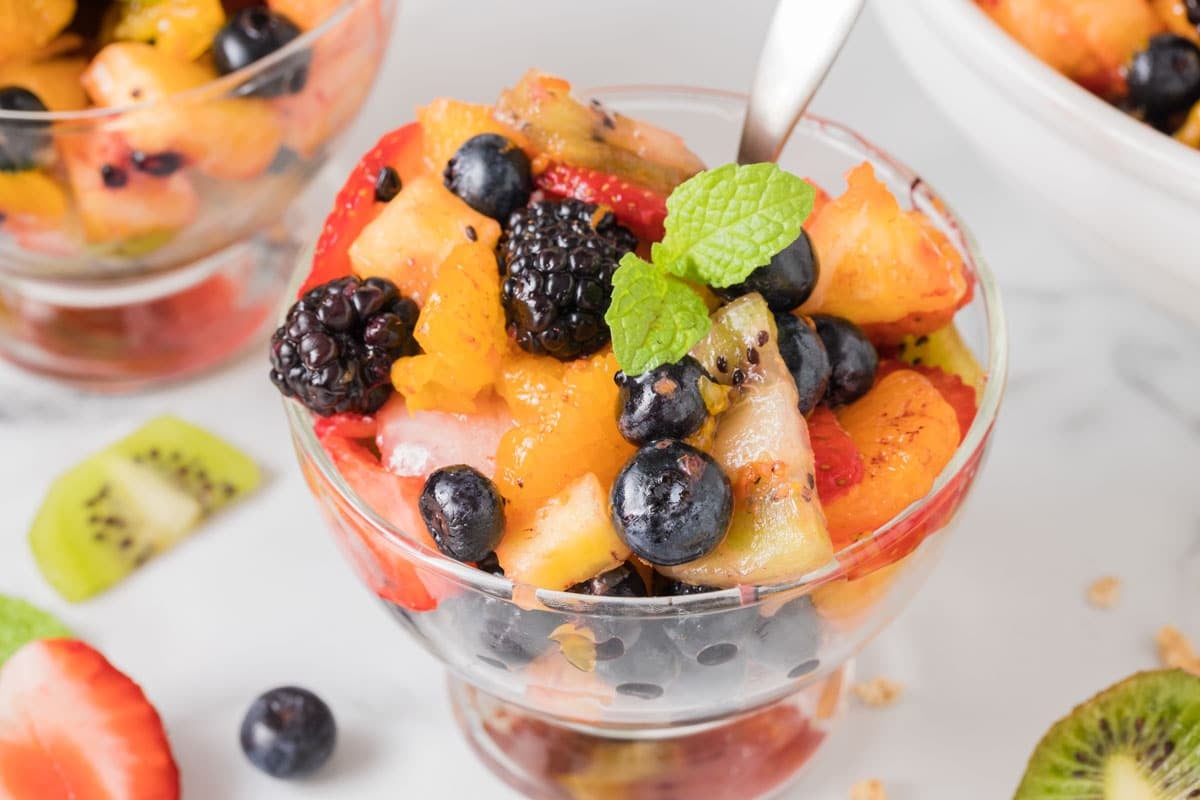 A glass bowl filled with assorted fresh fruit including blueberries, blackberries, kiwi, strawberries, and peaches, garnished with a mint leaf. A spoon rests in the bowl.