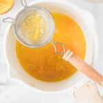 A small sieve with seeds and pulp rests over a pot of strained citrus juice on a white surface, surrounded by squeezed citrus halves.