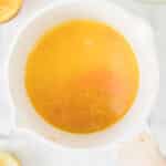 A white saucepan containing orange-yellow liquid and zest sits on a white countertop surrounded by citrus halves and a strainer.