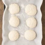 Six round pieces of raw dough sit on a parchment-lined baking sheet, placed on a wooden surface.