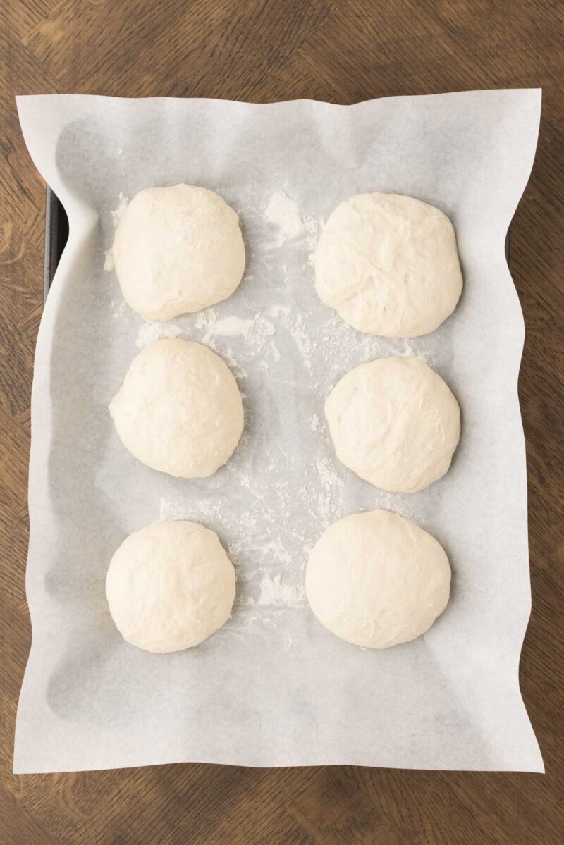 Six round pieces of raw dough sit on a parchment-lined baking sheet, placed on a wooden surface.