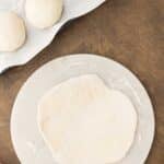 A flat, rolled-out dough rests on a marble surface next to three dough balls on a parchment-lined tray, with a rolling pin and pink cloth nearby.