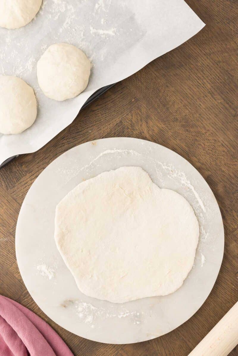 A flat, rolled-out dough rests on a marble surface next to three dough balls on a parchment-lined tray, with a rolling pin and pink cloth nearby.