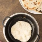 A round piece of dough is cooking in a black skillet, with a plate of cooked flatbreads on the side, all on a wooden surface.