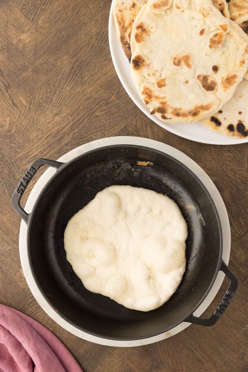 A round piece of dough is cooking in a black skillet, with a plate of cooked flatbreads on the side, all on a wooden surface.