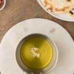 A saucepan with olive oil and minced garlic sits on a white board; nearby are a plate of flatbread and a bowl of chopped herbs.