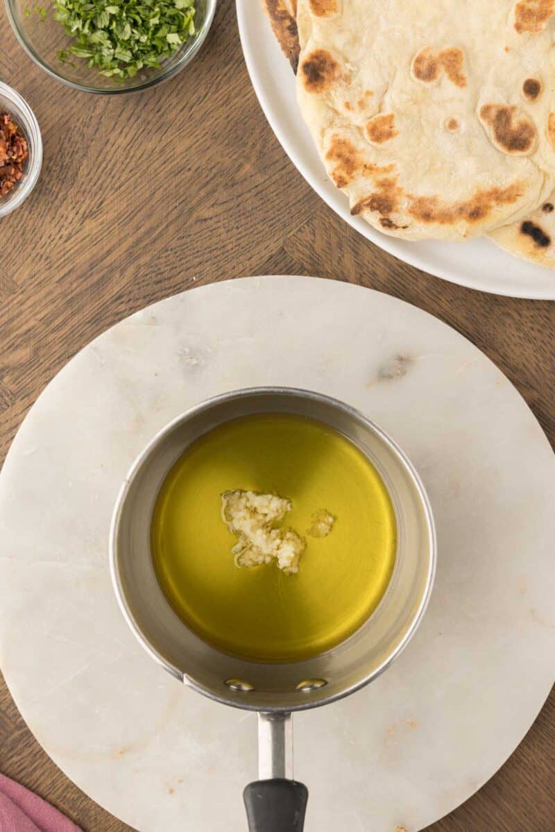 A saucepan with olive oil and minced garlic sits on a white board; nearby are a plate of flatbread and a bowl of chopped herbs.