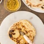 A hand brushes melted garlic butter onto a piece of cooked naan bread on a marble surface, with more naan and ingredients visible nearby.