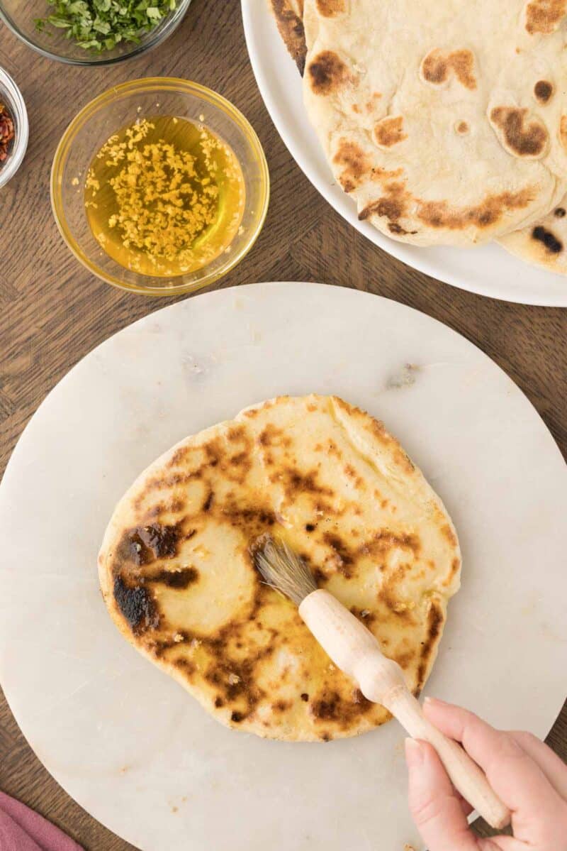 A hand brushes melted garlic butter onto a piece of cooked naan bread on a marble surface, with more naan and ingredients visible nearby.