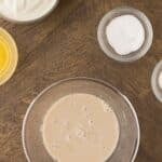 Overhead view of bowls containing yeast mixture, flour, salt, sugar, melted butter, and yogurt on a wooden table.