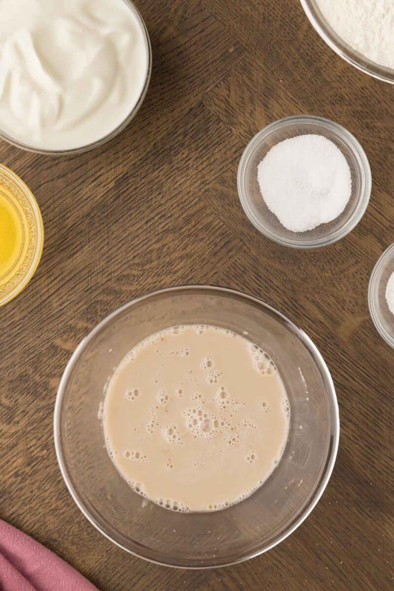 Overhead view of bowls containing yeast mixture, flour, salt, sugar, melted butter, and yogurt on a wooden table.