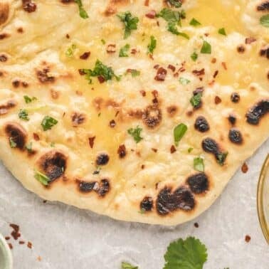 A piece of garlic naan topped with herbs and red pepper flakes sits on parchment paper, with bowls of seasonings and melted butter nearby.