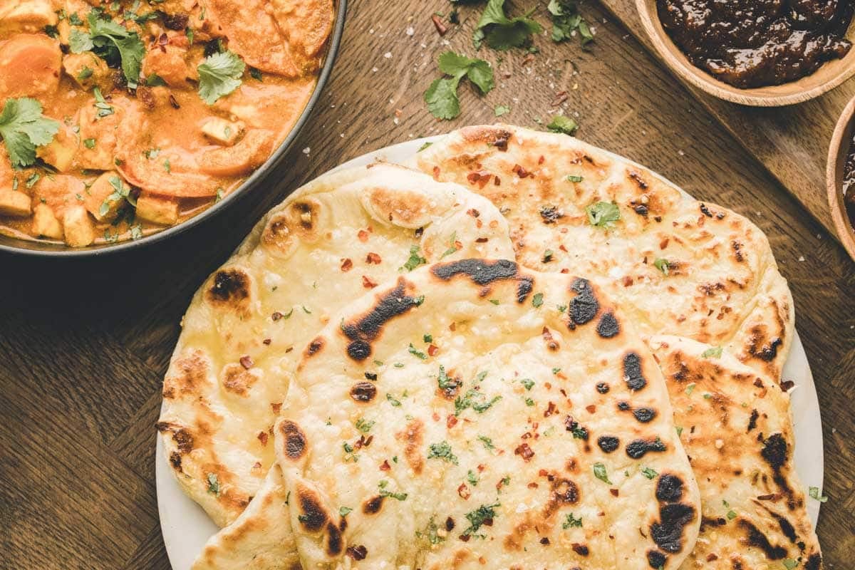 A plate of naan bread garnished with herbs, next to a bowl of curry with paneer and two small bowls of chutney on a wooden table.
