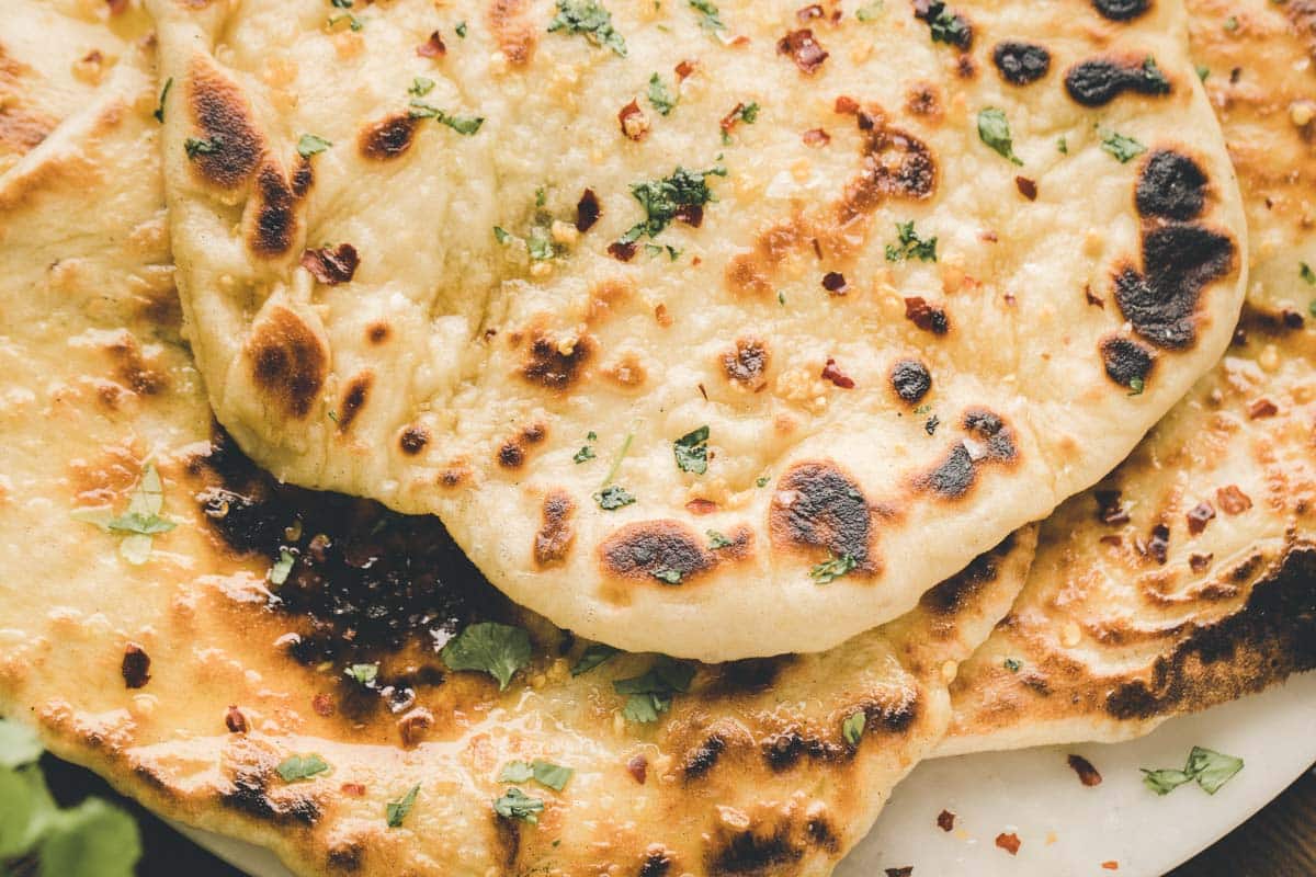 Close-up of several pieces of naan bread, garnished with chopped herbs and red chili flakes, showing toasted spots and a soft texture.