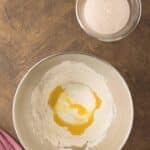 A mixing bowl with flour, yogurt, and oil, and a smaller bowl with foamy yeast mixture on a wooden surface next to a pink cloth.