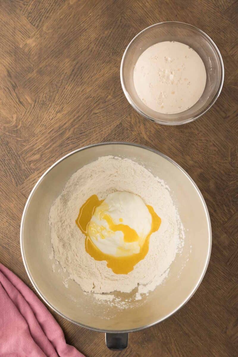 A mixing bowl with flour, yogurt, and oil, and a smaller bowl with foamy yeast mixture on a wooden surface next to a pink cloth.