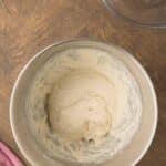 A metal mixing bowl with bread dough inside sits on a wooden surface next to a pink cloth and an empty glass bowl.