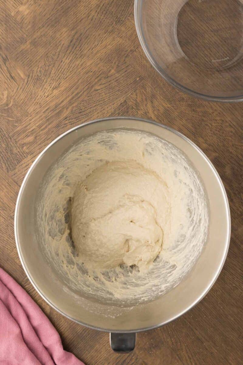 A metal mixing bowl with bread dough inside sits on a wooden surface next to a pink cloth and an empty glass bowl.