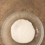 A ball of dough rests in a clear glass bowl on a wooden surface, with part of a pink cloth visible in the corner.