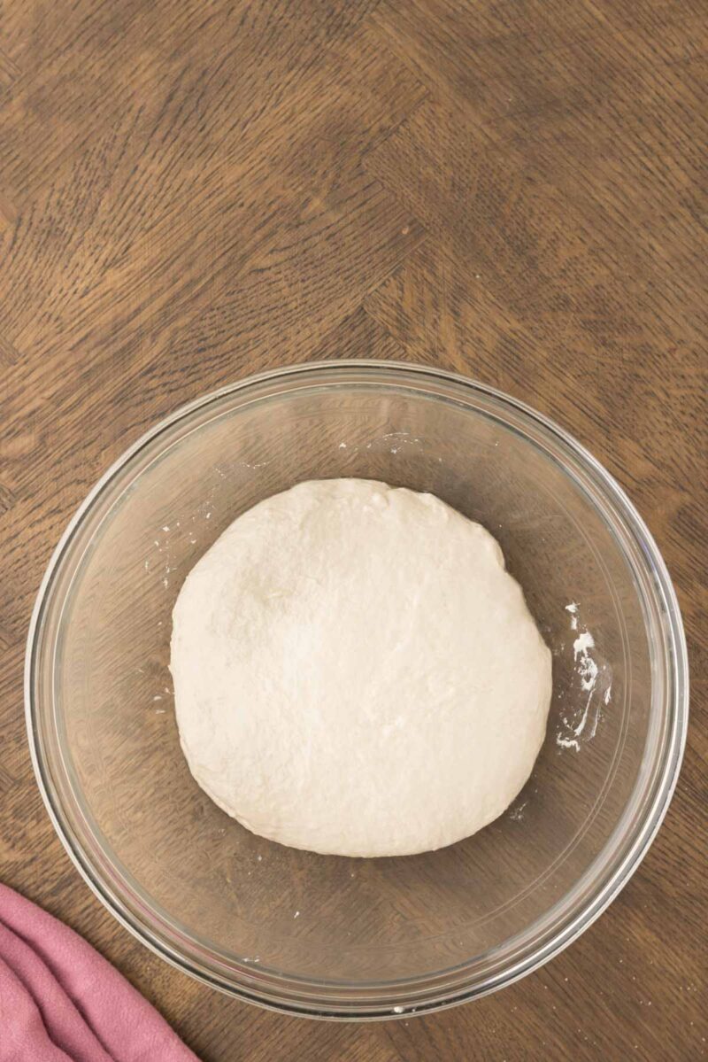 A ball of dough rests in a clear glass bowl on a wooden surface, with part of a pink cloth visible in the corner.