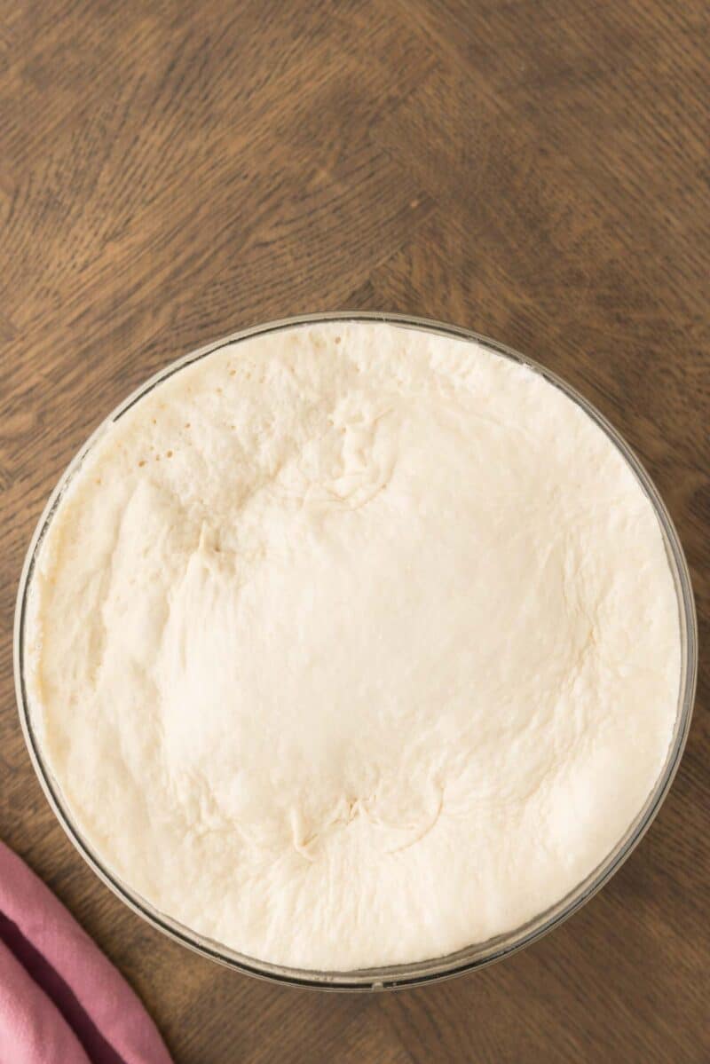 A glass bowl filled with risen pizza dough sits on a wooden surface, with a pink cloth partially visible in the corner.
