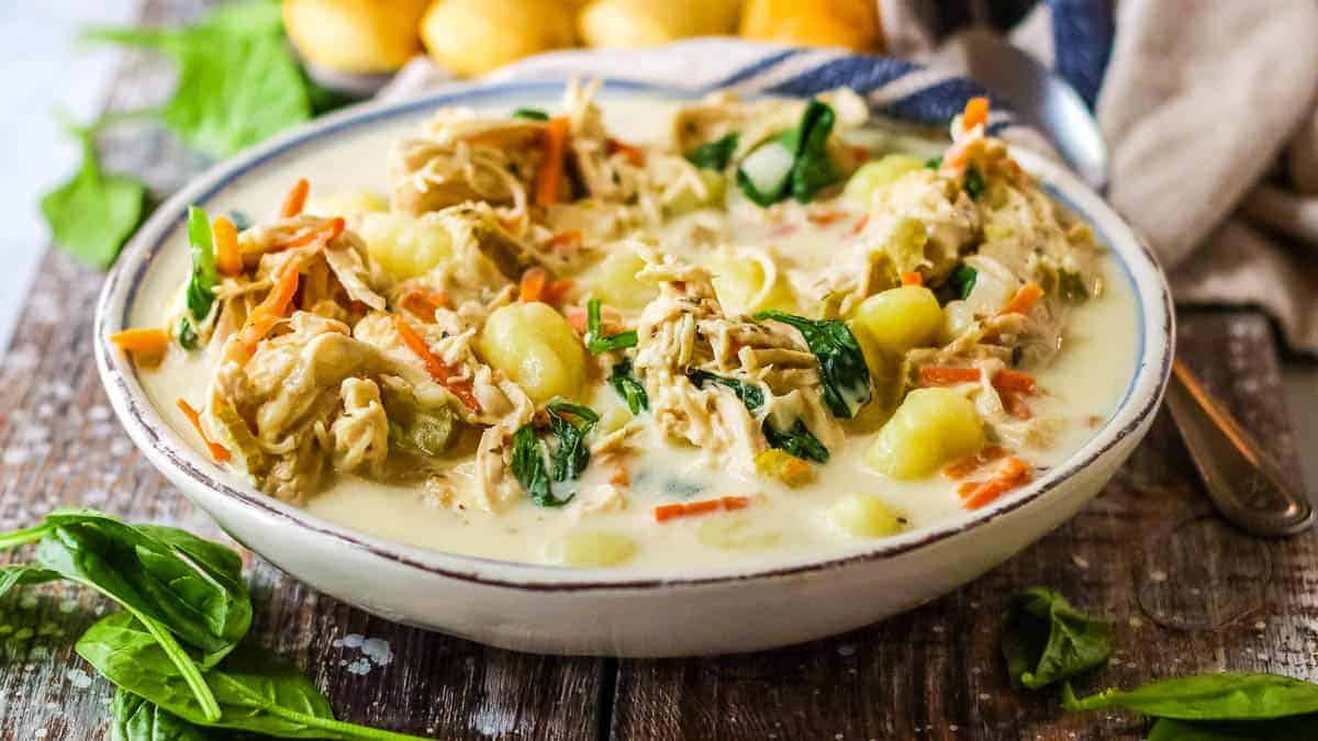 A bowl of creamy soup featuring saucy gnocchi, shredded chicken, spinach, and carrots sits on a wooden table with fresh spinach leaves and bread rolls in the background.