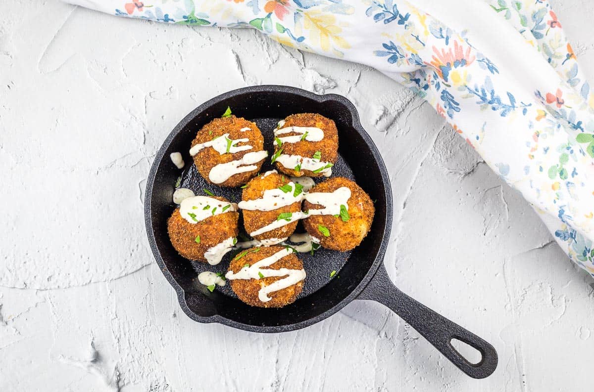 Round breaded patties make delicious bite-sized treats, topped with a drizzle of white sauce and chopped herbs, served in a small black skillet on a light textured surface next to a floral cloth.