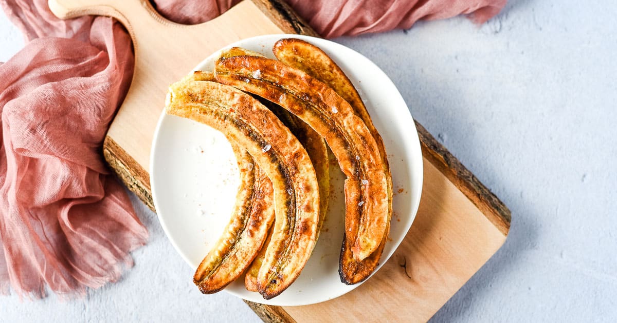 Three baked bananas with browned tops on a white plate, placed on a wooden board with a pink cloth in the background, showcase the warmth and flavor found in comforting cinnamon recipes.