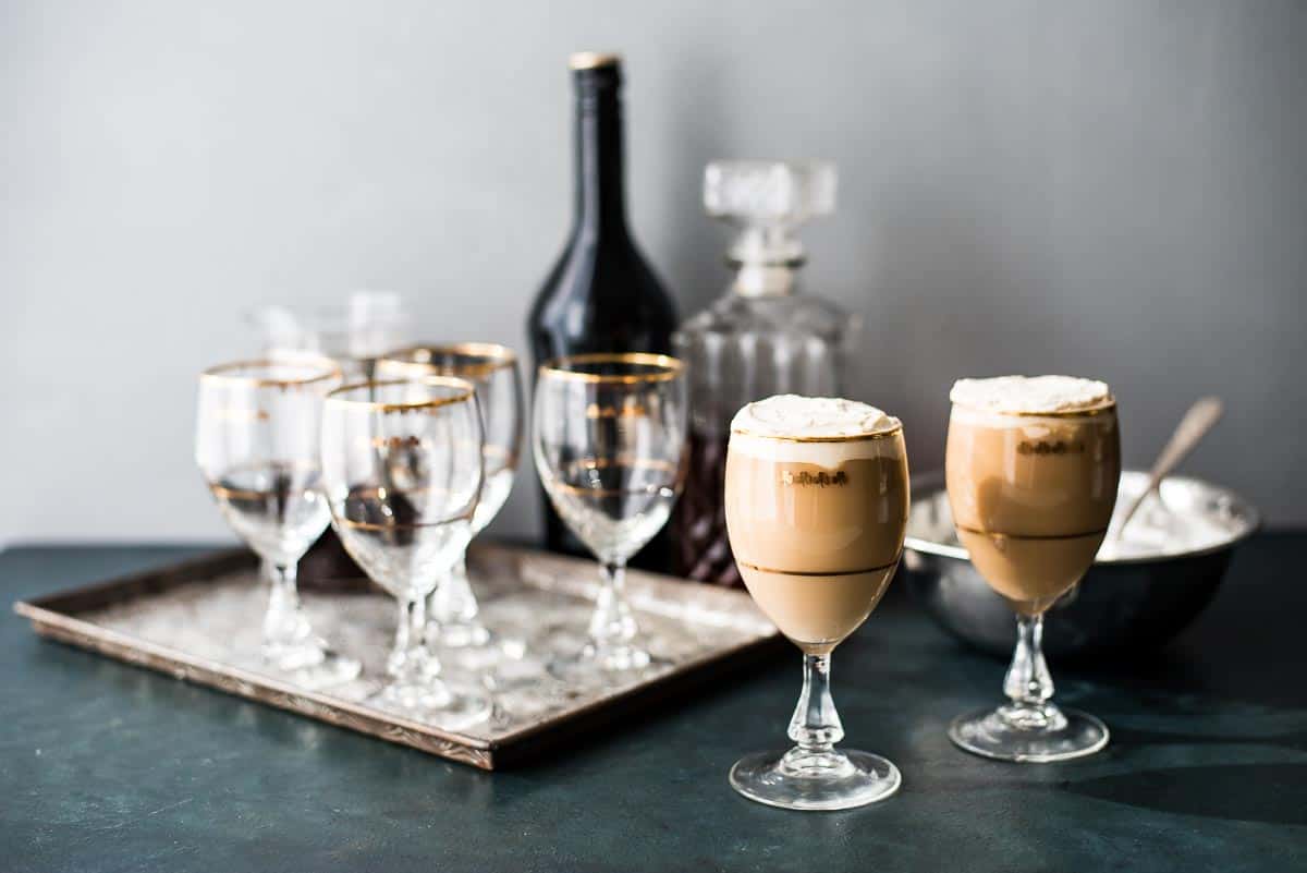 Two glasses of coffee with whipped cream are in the foreground, offering inspiring coffee ideas, with empty gold-rimmed glasses, a bottle, and a decanter on a tray in the background.