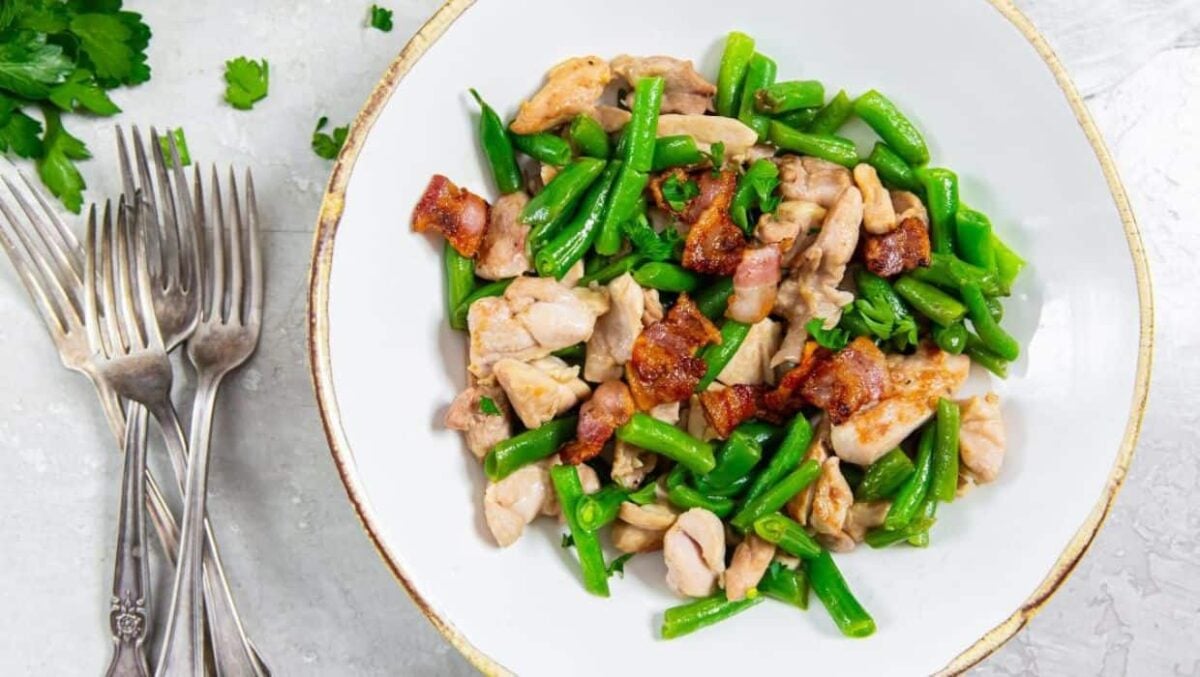 A white bowl filled with cooked green beans, pieces of chicken, and crispy bacon pieces sits on a light surface next to a small pile of forks and parsley—a perfect example of hearty entrees.