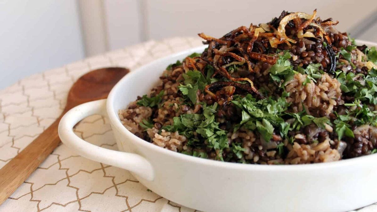 A white bowl filled with hearty lentil and rice pilaf, topped with chopped cilantro and crispy fried onions, sits on a patterned cloth with a wooden spoon beside it—a perfect choice for those who love flavorful entrees.