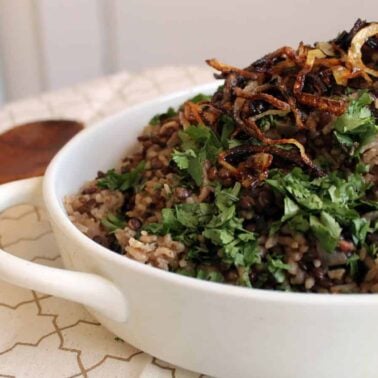 A white bowl filled with hearty lentil and rice pilaf, topped with chopped cilantro and crispy fried onions, sits on a patterned cloth with a wooden spoon beside it—a perfect choice for those who love flavorful entrees.