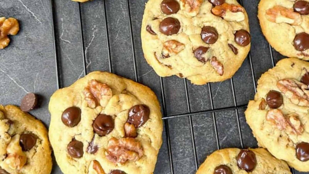 Chocolate chip cookies with crunchy walnut pieces cooling on a black wire rack, with a few chocolate chips and walnut pieces scattered nearby.