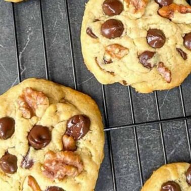 Chocolate chip cookies with crunchy walnut pieces cooling on a black wire rack, with a few chocolate chips and walnut pieces scattered nearby.