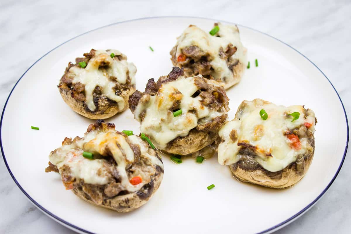 Five stuffed mushrooms topped with melted cheese and garnished with chopped chives are arranged on a white plate with a blue rim, making them perfect as savory snacks or baked appetizers for any gathering.