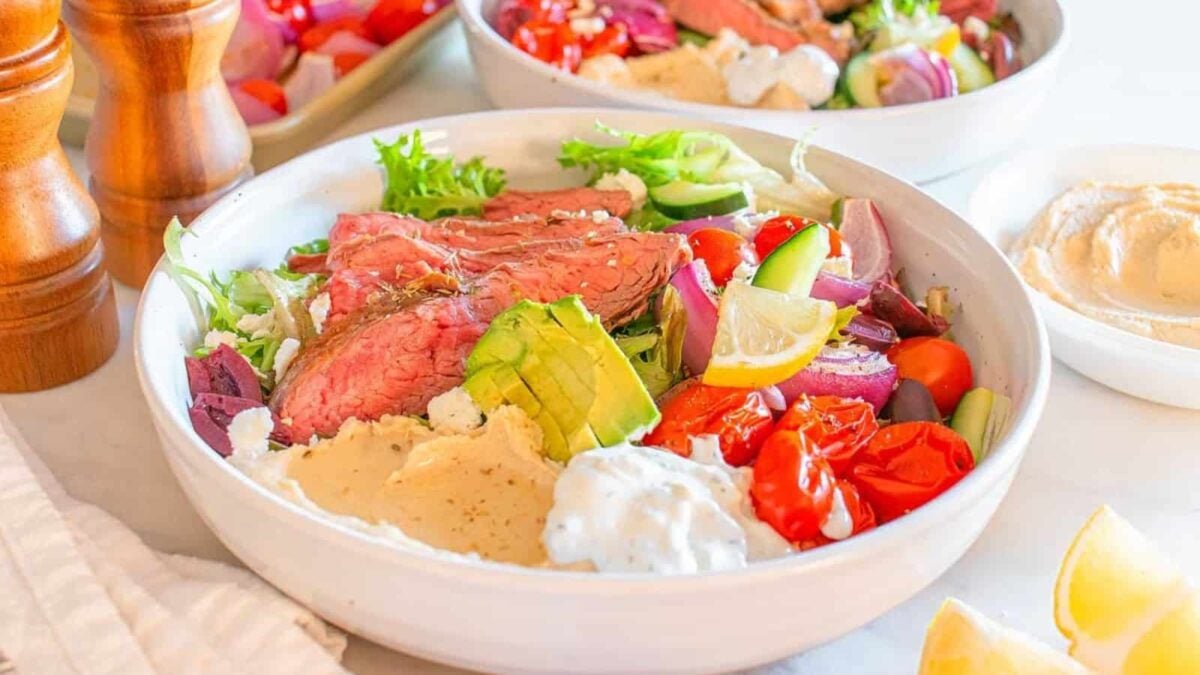 A bowl filled with sliced steak, mixed greens, avocado, tomatoes, cucumber, red onion, olives, hummus, tzatziki, lemon wedges, and feta cheese—a perfect gluten free beef dinner idea.