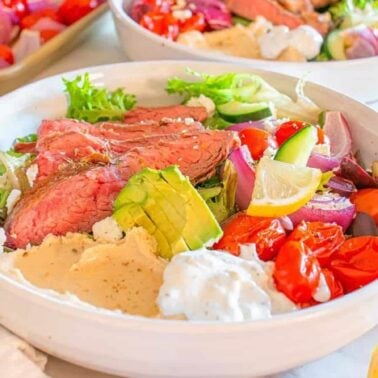 A bowl filled with sliced steak, mixed greens, avocado, tomatoes, cucumber, red onion, olives, hummus, tzatziki, lemon wedges, and feta cheese—a perfect gluten free beef dinner idea.