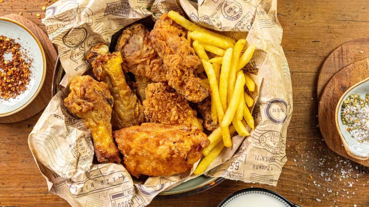 A basket lined with printed paper holds crispy fried chicken pieces and French fries, placed on a wooden table with small bowls of seasoning on the side.