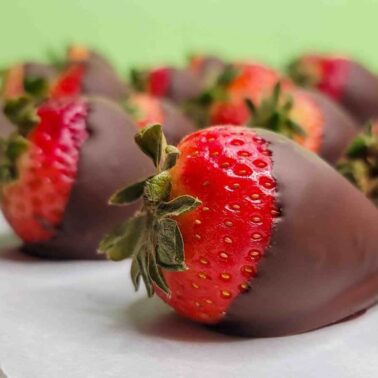 A close-up of a fresh strawberry partially dipped in chocolate, resting on parchment paper with more chocolate-covered strawberries in the backgroundโperfect vegetarian desserts for every occasion.