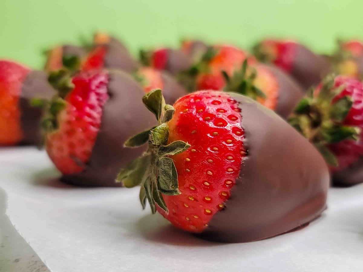 A close-up of a fresh strawberry partially dipped in chocolate, resting on parchment paper with more chocolate-covered strawberries in the background—perfect vegetarian desserts for every occasion.