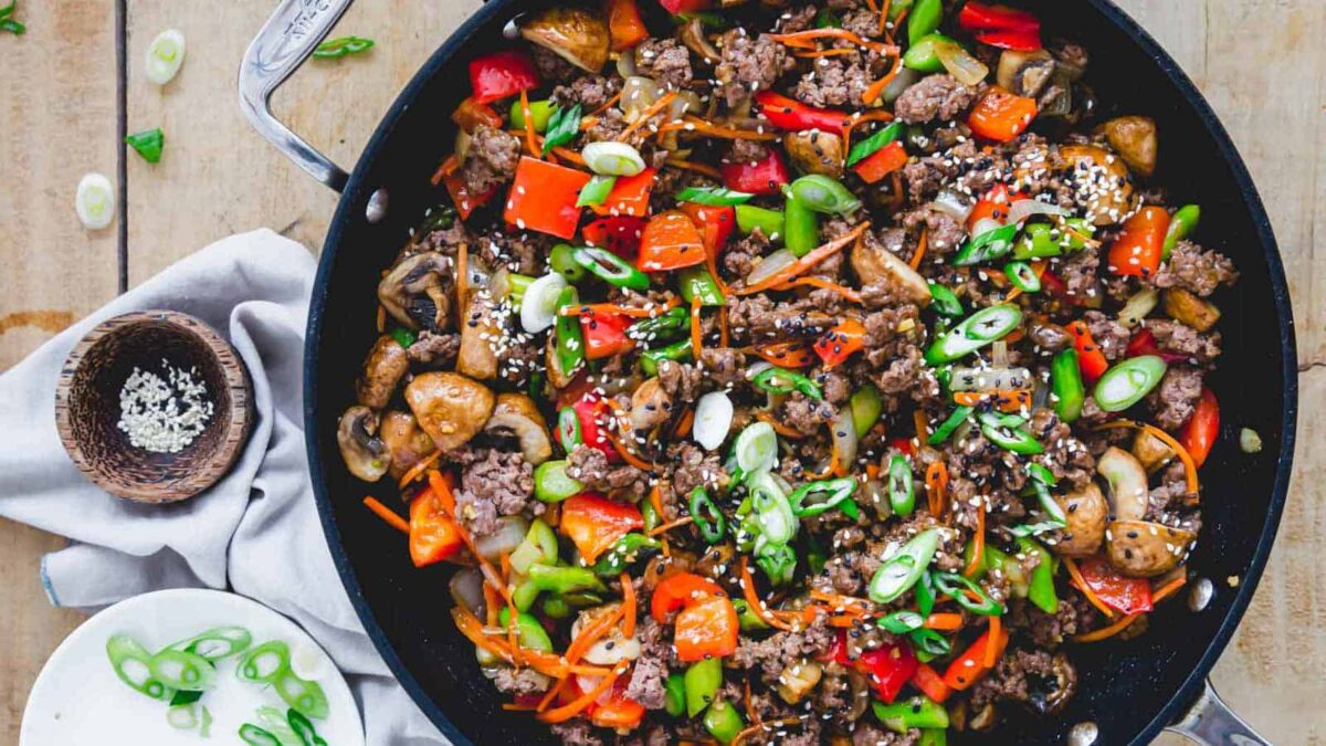 A skillet filled with hearty protein from ground beef, bell peppers, mushrooms, carrots, and green onions, garnished with sesame seeds. A bowl of sesame seeds and a plate of sliced green onions are nearby for this wholesome, gluten free meal.