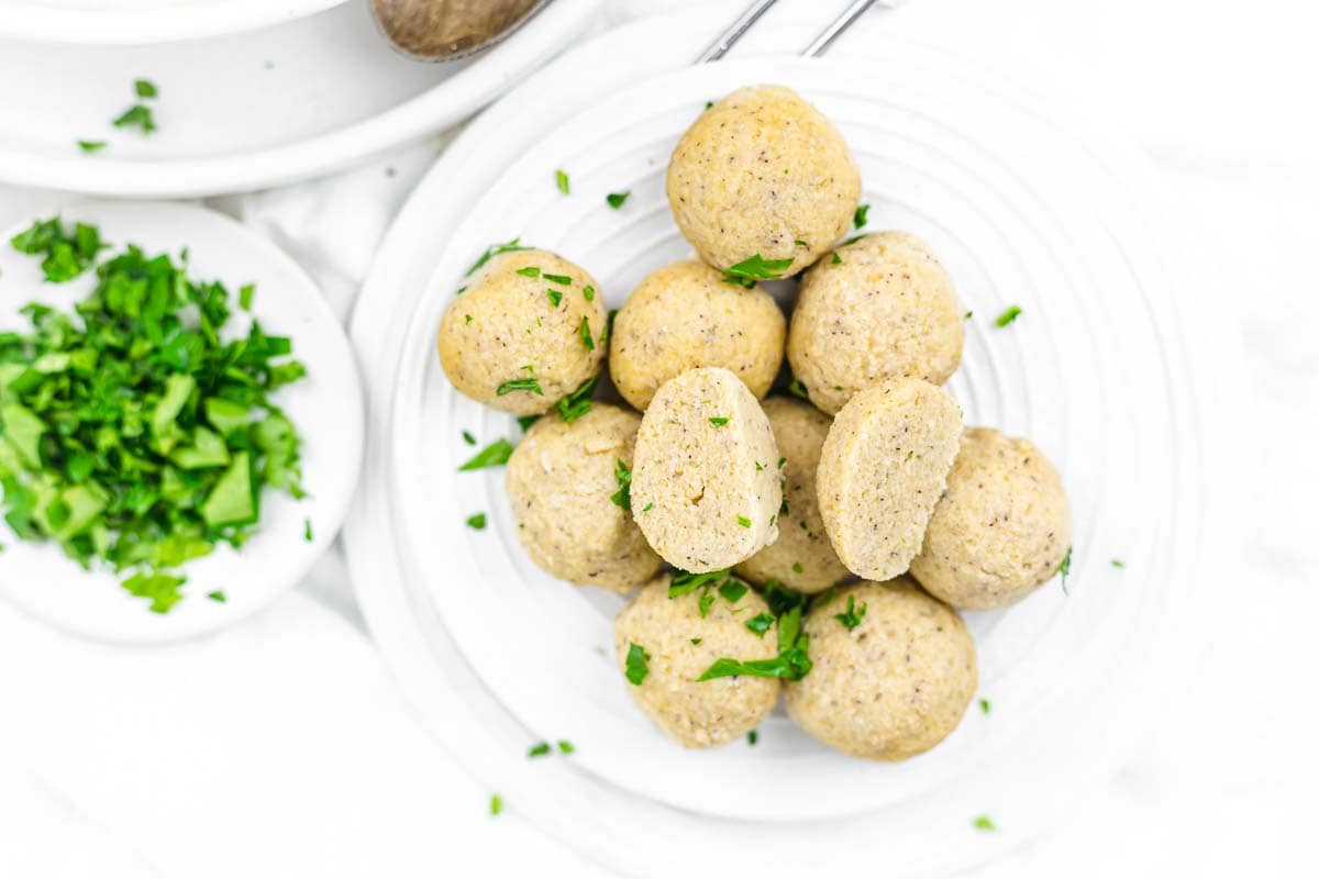 A white plate with several flavorful dumplings, shaped into round matzo balls and sprinkled with chopped fresh herbs, sits next to a small dish of more chopped herbs.