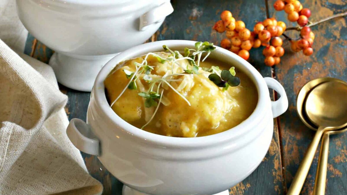 A white soup bowl filled with creamy yellow soup and soft dumplings, garnished with microgreens, sits on a rustic wooden table beside a gold spoon and orange berries.