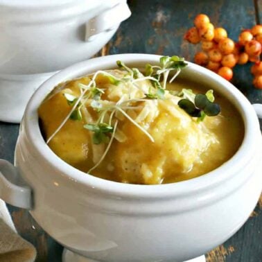 A white soup bowl filled with creamy yellow soup and soft dumplings, garnished with microgreens, sits on a rustic wooden table beside a gold spoon and orange berries.