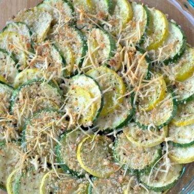 A glass dish filled with fresh baked yellow squash and zucchini slices, topped with savory breadcrumbs and shredded cheese, sits on a wooden surface.