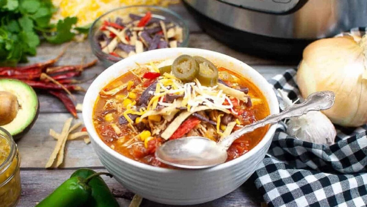 A bowl of tortilla soup topped with shredded cheese, tortilla strips, and jalapeños sits on a table with a spoon, surrounded by fresh ingredients and an Instant Pot—perfect inspiration for fun tortilla ideas and delicious tortilla recipes.