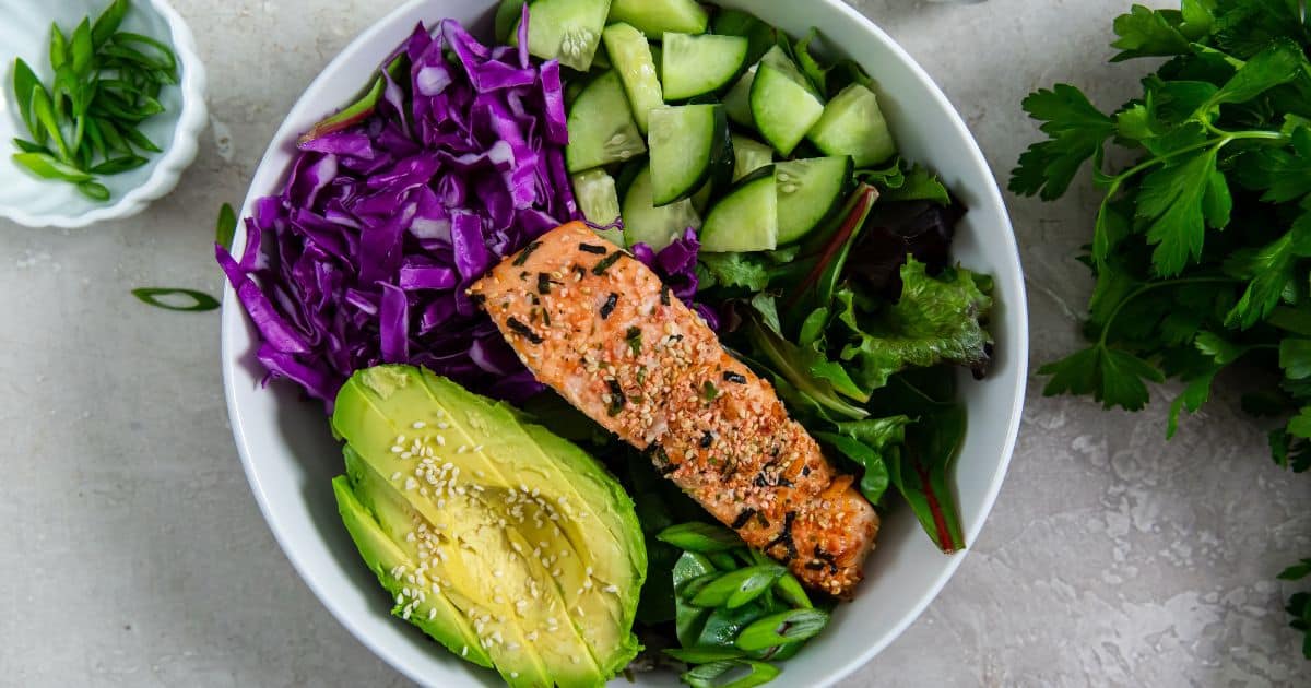 A bowl with salmon, sliced avocado, shredded purple cabbage, crunchy cucumber chunks, mixed greens, and green onions, all garnished with nutty sesame seeds.