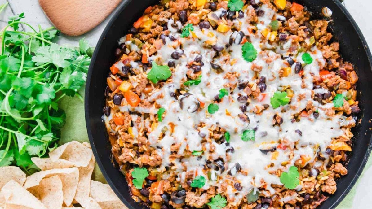 A skillet filled with cooked ground beef, black beans, diced vegetables, and melted cheese, garnished with cilantro—this easy dinner is perfect for anyone seeking satisfying meals. Served alongside tortilla chips and fresh cilantro.
